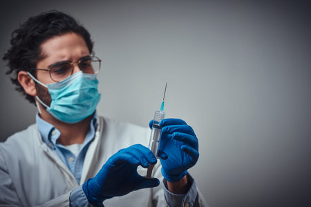 Serious medic in protective mask and gloves is holding a syringe with a medication.
