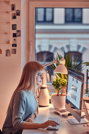 Focused blond student doing her homework while sitting next to the table and using computer.の写真素材