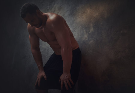 In the dark room tired shirtless man is leaning to the wall.