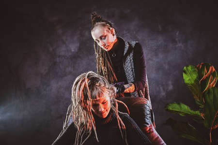 Woman with long dreadlocks is making dreads for blond girl at dark room.の写真素材