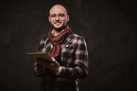 Succesful and smiling stylish bald businessman posing for camera in a dark studio wearing checkered wool jacket, glasses and ruby patterned scarf while holding tabletの写真素材