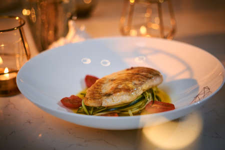 Delicious fillet of grilled and oven baked codfish or coalfish served with thin sliced vegetables and oil sauce, on a white porcelain dish next to golden candles in a warm light of a restaurant, close up view from the topの写真素材
