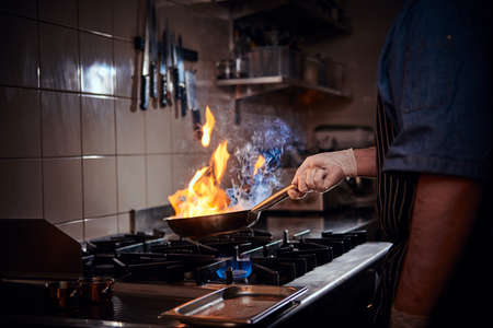 Professional chef wearing gloves and apron preparing stir-fry flambe, creating flames, holding a pan with one hand in a dark restaurant kitchenの写真素材