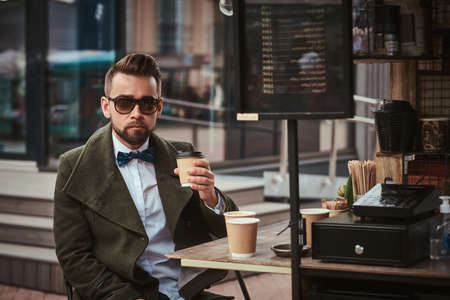 Stylish and elegant young male customer sitting outdoors next to coffee making gear in a mobile coffee shop in a city emporium, wearing green wool coat, white shirt and bow tie, taking a sip of coffee of the paper cup in the morning looking calm and seriousの写真素材
