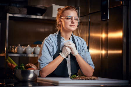 European beautiful female chef sitting in a dark restaurant kitchen, wearing glasses, gloves, apron and denim shirt, holding a knife against cutting board near sliced vegetables, looking patient.の写真素材