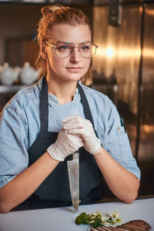 European beautiful female chef sitting in a dark restaurant kitchen, wearing glasses, gloves, apron and denim shirt, holding a knife against cutting board near sliced vegetables, looking patient.の写真素材