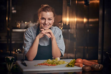 European beautiful female chef standing in a dark kitchen next to cutting board with vegetables on it, wearing apron and denim shirt, posing for the camera, holding hands under the chin, reality show lookの写真素材
