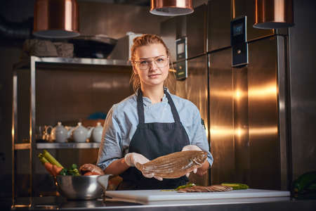 Beautiful and stylish woman chef standing in a dark restaurant kitchen next to cutting board with vegetables on it, holding a big fish and wearing apron and denim shirt, posing for the camera, cooking show lookの写真素材