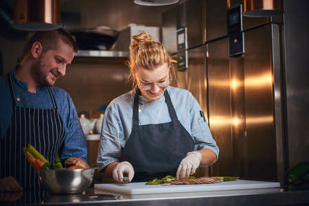 Smiling male and female chefs standing in a dark kitchen next to cutting board with vegetables on it, wearing aprons and denim shirts, posing for the camera, cooking show style, looking interested and patientの写真素材