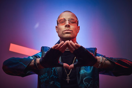 Creative, daring, tattooed, bald male rocker model posing in a studio for the photoshoot wearing fashionable designer black leather jacket, black shirt, red glasses, and jewelery, showing a triangle with armsの写真素材