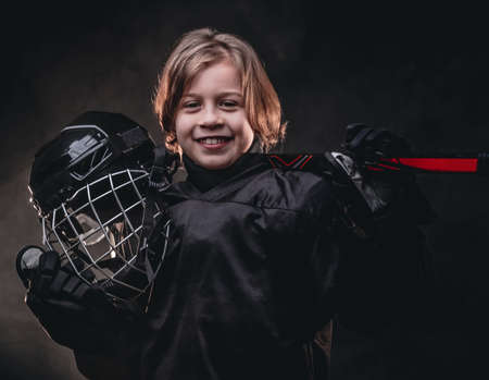 Young blonde sporty boy, ice hockey player, posing in a dark studio for a photoshoot, wearing an ice-skating uniform while holding his helmet, hockey stick and smiling, looking confident and braveの写真素材