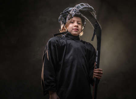 Young, blonde, confident, sporty boy, ice hockey player, posing in a dark studio for a photoshoot, wearing an ice-skating uniform while wearing his helmet, holding a hockey stick and looking calmの写真素材