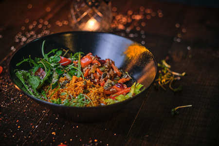 Healthy, vegetarian plate of meal which includes cooked vegetarian pilaf served with healthy mix of ruccola leafs, bulgarian pepper, thin sliced vegetables, cherry tomatoes and spices, on a black porcelain dish, decorated wth spices on a wooden table in a warm light of a restaurant, close up view from the sideの写真素材