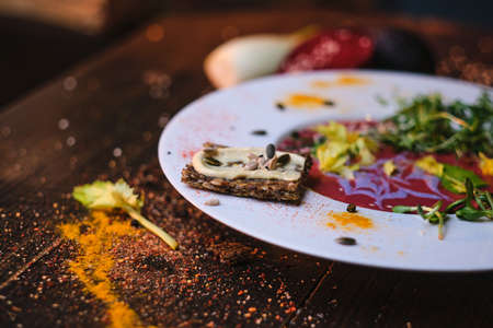 Healthy, vegetarian appetizer of beetroot sauce served with vegan hummus spread on a thin sliced grain cracker, near mixed green sprouts on a white porcelain dish next to a beetroot and a root-crop, decorated with spices on a wooden table in a warm light of a restaurant, close up view from the sideの写真素材