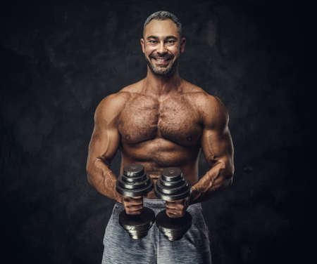 Strong, adult, fit muscular caucasian man coach posing for a photoshoot in a dark studio under the spotlight wearing grey sportswear, showing his muscles and putting up a dumbbells looking joyfulの写真素材