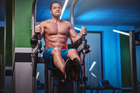 Powerful athletic bodybuilder doing excersise on a parallel bars, pumping his abs and lifting his...