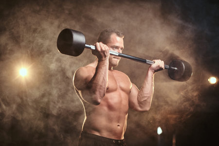 Fit mucular bodybuilder doing weightlifts with barbell in a dark gym surrounded by smoke looking focusedの写真素材