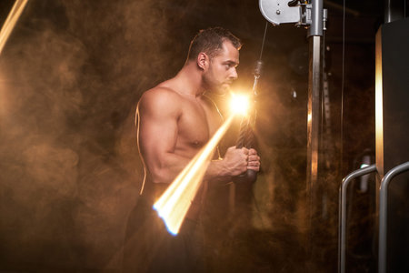 Strong shirtless male athlete concentrated on an excersise on a hand pull machine, pumping up his...