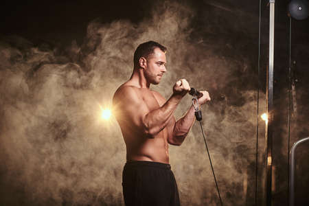 Powerful adult bodybuilder pumping up his biceps on a hand pull machine in a dark gym under the spotlights surrounded by smoke looking calmの写真素材