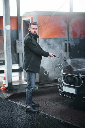 Well-dressed young adult man washing his black car outdoors in a carwash under high pressured water...