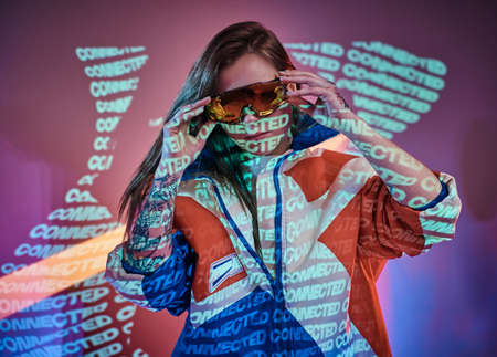 Stylish and futuristic young model posing in neon studio lights of neon text projection over pink background, wearing bright racing blazer and holographic sunglasses. Cyberpunk conceptの写真素材