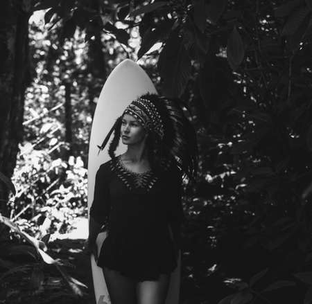 Beautiful and confident semi-nude female wearing american indian traditional headdress, while posing in the forest and standing in front of a surfer board. Black and white close-up photoの写真素材