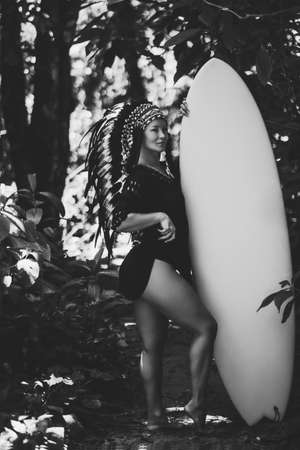 Sexy and seductive half-nude female wearing american indian traditional headdress, while posing in the forest and holding a surfer board. Black and white photoの写真素材