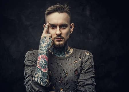 Inked young man wearing grey ripped jumper posing looking thoughtful, isolated on a grey background in a dark studio. Close up photoの写真素材