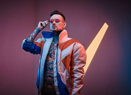Extravagant male model tattoo artist posing in a neon studio wearing racer coat on a half-naked body, fire-shaped sunglasses and tattooed in a japanese irezumi style, looking self-assured.の写真素材