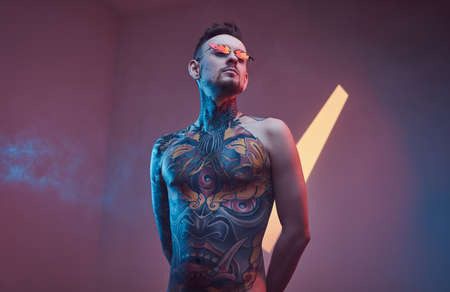 Cool male model posing in a neon studio with a half-naked body wearing fire-shaped sunglasses and...