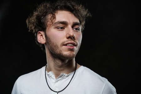 Curly and hansome caucasian man standing in a dark studio on a grey background, wearing casual white shirt looking interestedの写真素材