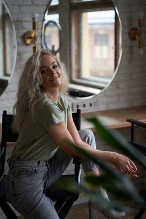 Beautiful and elegant young woman with blonde curly hair sitting on a make up chair in a bright room...