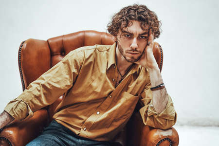 Portrait of a serious young adult guy with curly hair, wearing luxury orange shirt while sitting on the leather chair, looking authoritativeの写真素材