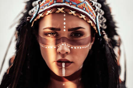 Beautiful and confident female wearing American indian traditional headdress, leather jacket and sunglasses while posing in a bright studio and looking seriousの写真素材