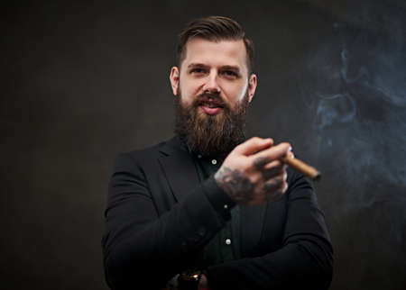 Elegantly dressed bearded male with tattoos on his neck and hand lights a cigar. Brutal young businessman smokes a cigar in a dark studio.の写真素材