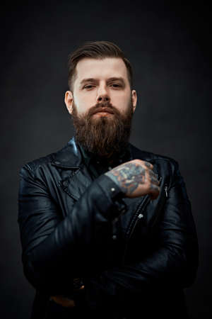 Cool bearded tattooed man in a leather jacket looking on the camera in a dark studioの写真素材