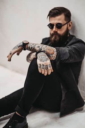 Stylish bearded guy with tattoos on his hands and neck sitting on a floor in a white studioの写真素材
