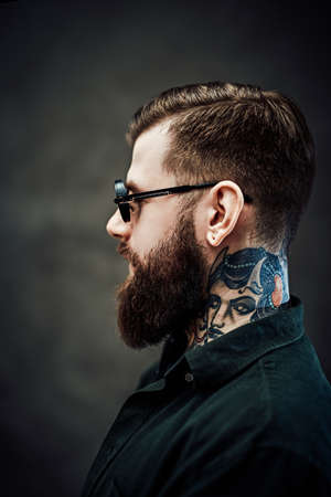Profile of a young bearded barber with tattoo on his neck in a dark studio