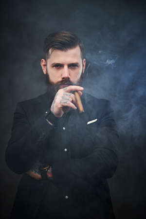 Elegantly dressed bearded businessman holds a cigar and looks at the camera in a smoky dark studioの写真素材