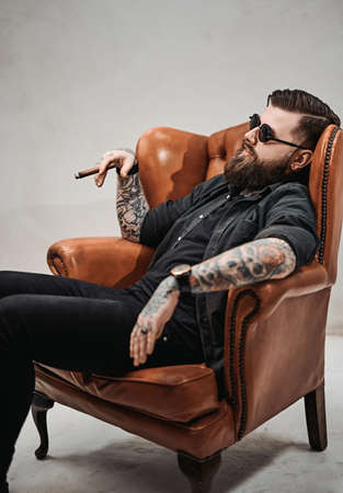 Cool tattooed bearded man smokes a cigar and relaxing on a vintage chair