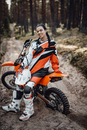 Beautiful young female racer sitting on her motocross bike on a trail of sand in the woods