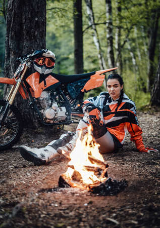Beautiful young female racer wearing motocross outfit, warming up sitting next to the bonfire