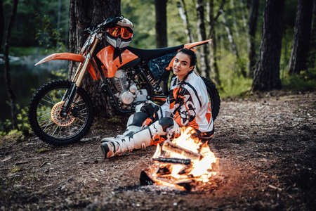 Beautiful young female racer wearing motocross outfit, warming up sitting next to the bonfireの写真素材