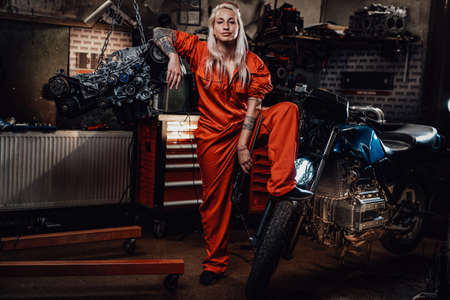 Blond female mechanic with tattooed hands in orange overalls stands next to the suspended flat engine and naked bike in garage or workshopの写真素材