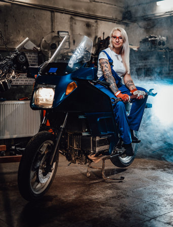 Young blond woman mechanic in work overalls hold big wrench while sitting on sportbike in garage or...