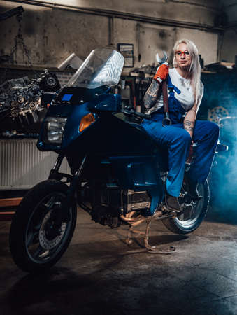 Young blond woman mechanic in work overalls hold big wrench while sitting on sportbike in garage or workshop, smiling and looking on cameraの写真素材