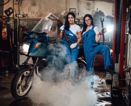 Two young female mechanic posing next to a sportbike in authentic workshop garage surrounded by...
