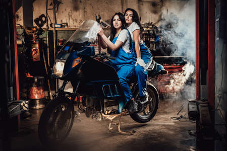Two beautiful female mechanic sitting on sportbike in authentic workshop garageの写真素材