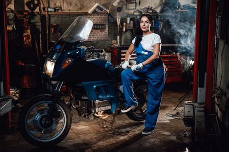 Beautiful brunette woman in blue overalls posing with big wrench while standing next to a sportbike in garage or workshopの写真素材