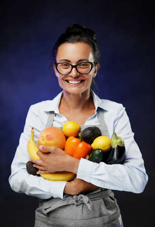 Attractive middle-aged woman in glasses and apron holds a lot of fresh vegetables and fruit, smiling and looking on a camera. Studio photo on a dark background with blue lightsの写真素材